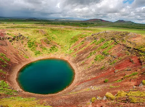 Kerid volcanic crater in Iceland with a deep blue-green lake surrounded by colorful red volcanic rock and green terrain.