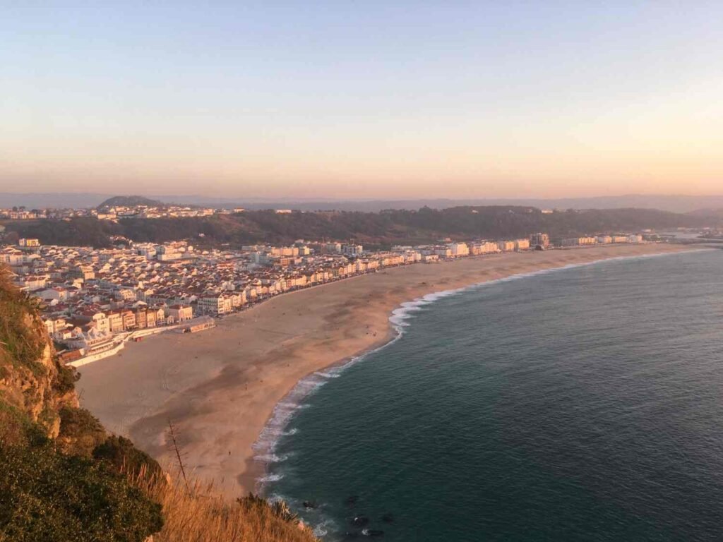 Wide sandy beach and coastal town of Nazaré, Portugal, viewed from a cliff at sunset.